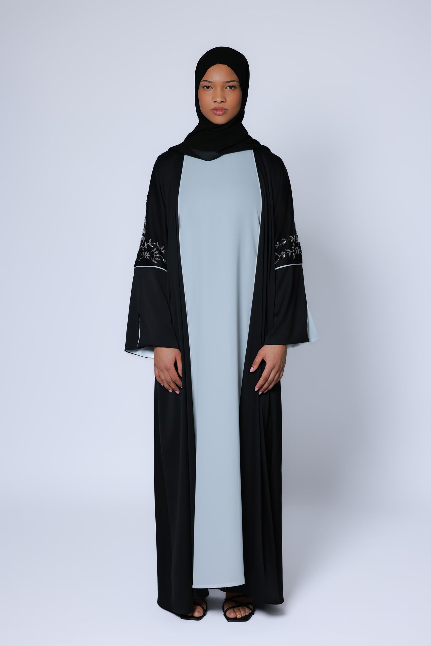Vooraanzicht van zwarte abaya met zilverkleurig bloemenborduurwerk op de mouwen, gedragen over een lichtblauwe onderjurk