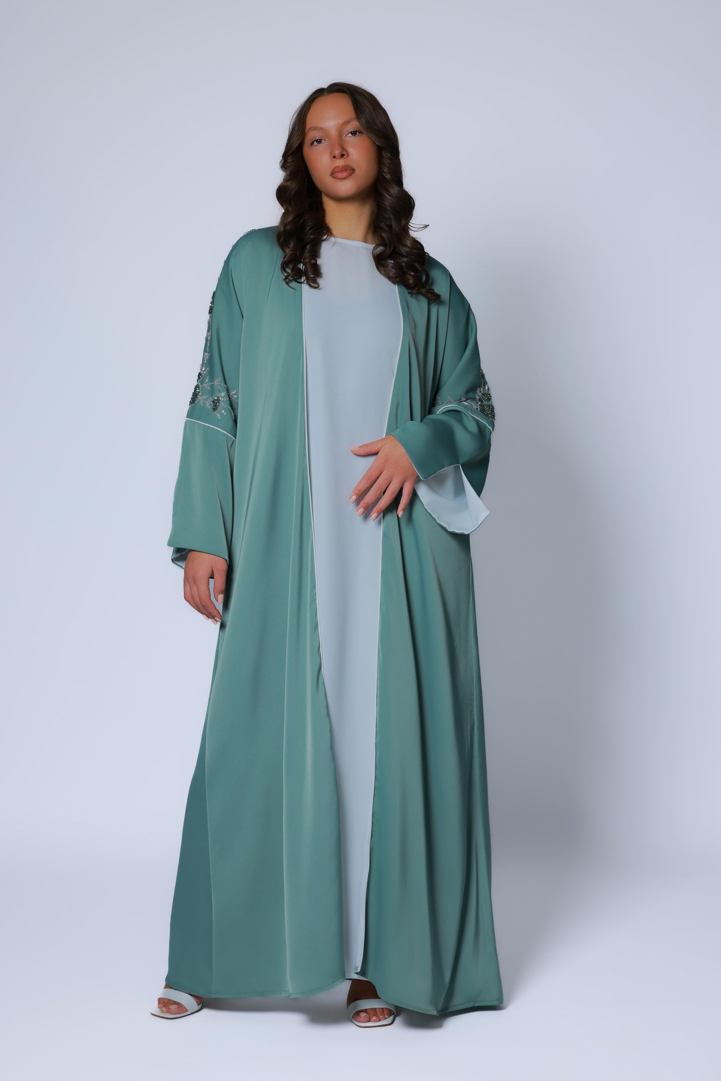 Vooraanzicht van een elegant vallende groenblauwe abaya met bloemenborduurwerk op de mouwen, gedragen met een lichtgrijze onderjurk
