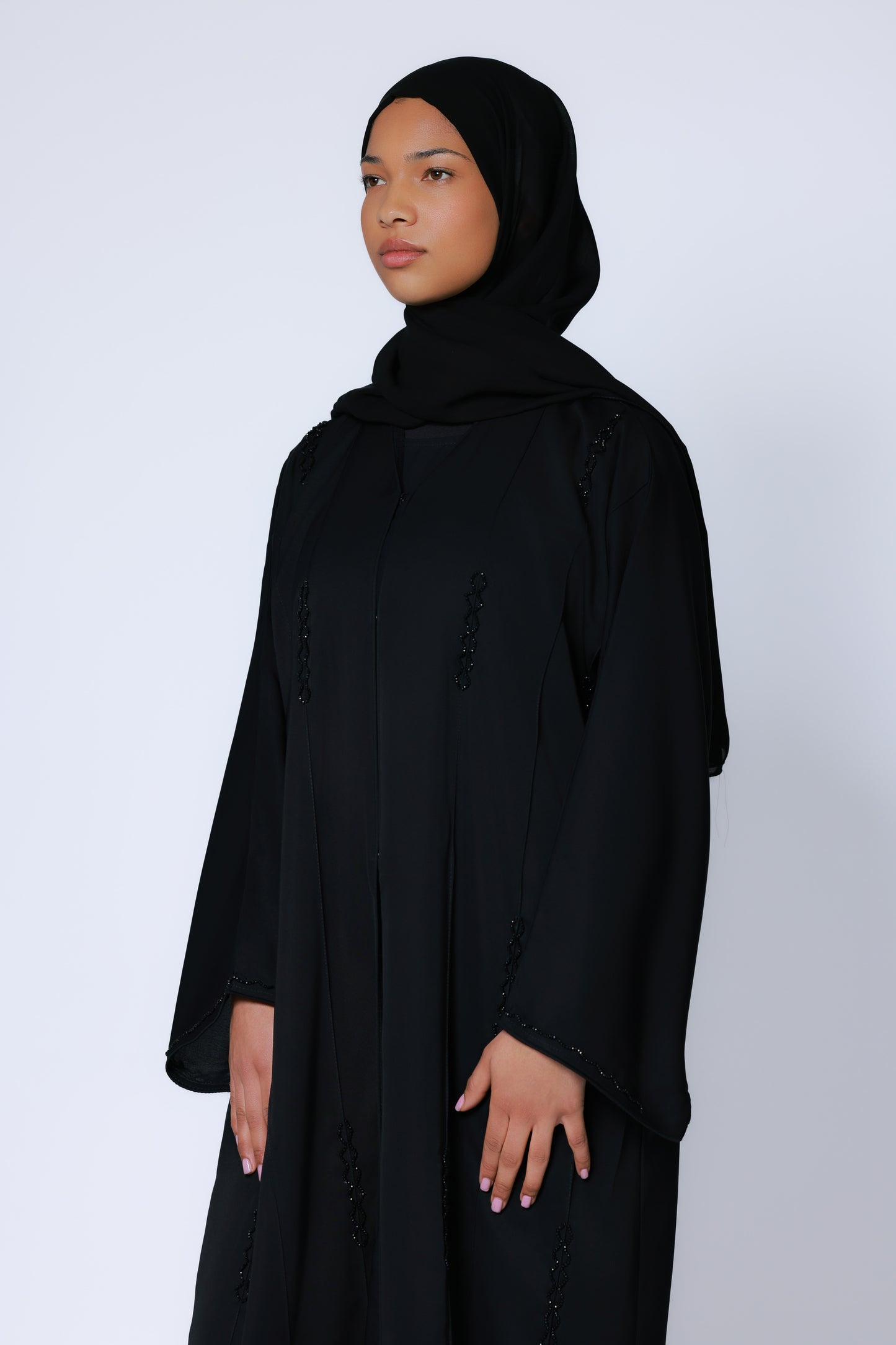 Model in zwarte abaya met hijab, staand tegen witte achtergrond, benadrukt vloeiende pasvorm
