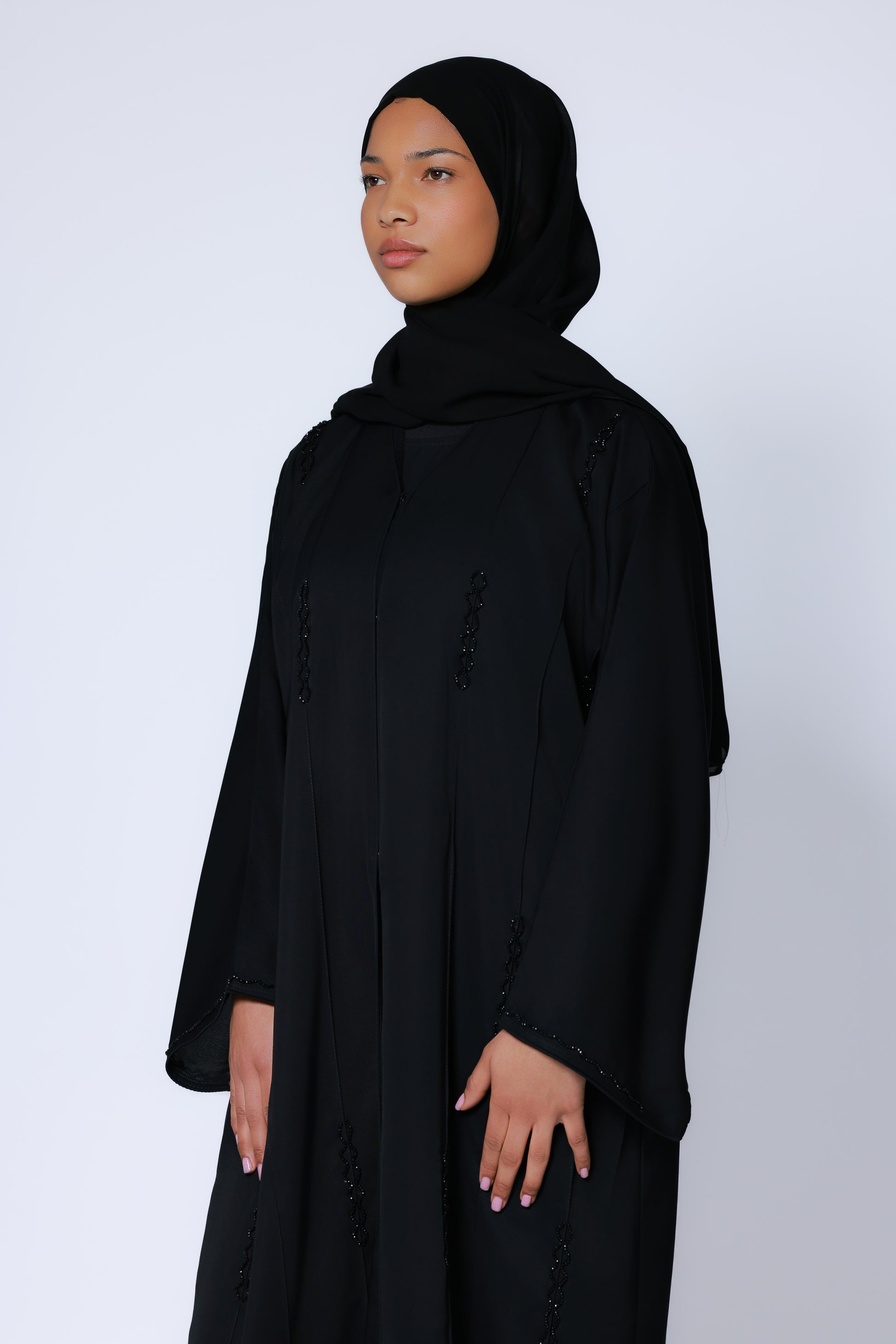 Model in zwarte abaya met hijab, staand tegen witte achtergrond, benadrukt vloeiende pasvorm