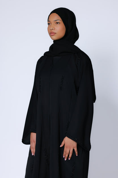 Model in zwarte abaya met hijab, staand tegen witte achtergrond, benadrukt vloeiende pasvorm